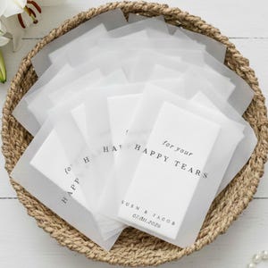 Peut inclure: Un panier en osier rempli de sachets en papier de soie blancs. Chaque sachet porte l'inscription "for your HAPPY TEARS", un nom et une date. Des fleurs blanches et des alliances dorées sont visibles en arrière-plan.