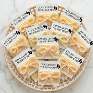 Puede incluir: Una cesta tejida llena de tarjetas de juego para baby shower. Cada tarjeta dice "CAN YOU GUESS THE BABY BUMP?" y está envuelta con hilo amarillo y atada con un lazo. Las tarjetas también presentan una huella.