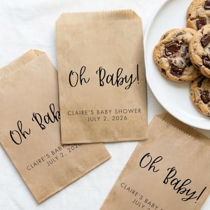 May include: Brown paper treat bags with the text "Oh Baby!" in black script. The bags are personalized with "Claire's Baby Shower July 2, 2026". Chocolate chip cookies are on a white plate next to the bags.