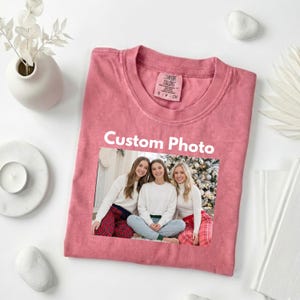 May include: A dusty rose Comfort Colors t-shirt with a custom photo print. The photo features three women in white sweaters and plaid pajama pants. The text "Custom Photo" is printed above the photo.