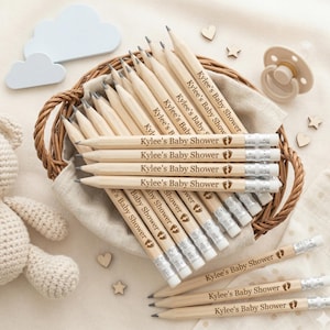 Puede incluir: Un conjunto de lápices de madera, cada uno grabado con "Kylee's Baby Shower" y un diseño de huella, se presenta en una cesta de mimbre. Los lápices tienen gomas de borrar plateadas y puntas de grafito. Estrellas, corazones, un chupete y una nube decoran el conjunto.