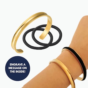 May include: Gold-colored bangle bracelet with a rounded design. The image also shows a black hair tie and a wrist wearing a gold bracelet with a black band. The text in the image says, "Engrave a message on the inside!"