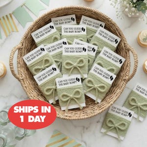 May include: A woven basket filled with baby shower game cards. Each card is wrapped with green twine and tied with a bow. The cards read "CAN YOU GUESS THE BABY BUMP?" and feature a footprint graphic.