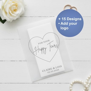 May include: A white wedding favour with the text "FOR YOUR Happy Tears" inside a heart shape. The favour is personalised with the names "CLAIRE & LUIS" and the date "02.05.2026". A blue circle with the text "+ 15 Designs • Add your logo" is also visible.