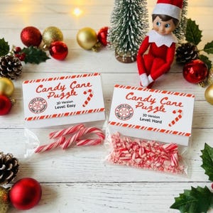 Puede incluir: Dos bolsas transparentes con rompecabezas de bastones de caramelo. Una contiene bastones enteros, la otra trozos triturados. Cada una tiene una etiqueta blanca con texto rojo: "Candy Cane Puzzle", "3D Version" y nivel de dificultad.