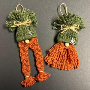 May include: Two handmade St. Patrick's Day gnome ornaments. Each gnome has a green yarn hat with a twine bow and a silver shamrock charm. One gnome has orange braided yarn legs, the other has an orange yarn beard. Both have a wooden button.