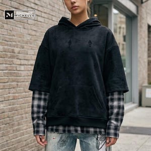May include: Black distressed hoodie with a layered plaid shirt underneath. The hoodie has a front pocket and a hood. The plaid shirt is black and white. The brand logo is in the top left corner.