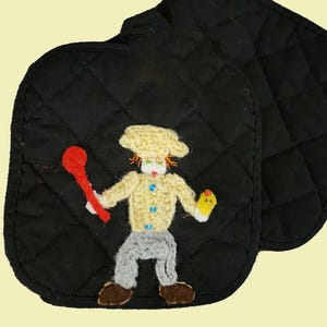May include: A black quilted pot holder featuring a crocheted applique of a chef figure. The chef wears a cream-colored hat and jacket with blue buttons, gray pants, and brown shoes, holding a red spoon in one hand and a yellow item in the other.