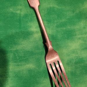 May include: A metallic fork with a long, slender handle featuring a rounded, spade-shaped end with a small engraved arch detail. The fork has four thin, pointed tines and a smooth, polished finish that reflects light across its surface.