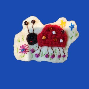 Puede incluir: Broche de insecto de fieltro hecho a mano con cuerpo rojo de ganchillo, cabeza negra y antenas marrones. La base de fieltro blanco presenta flores bordadas, hierba y una estrella amarilla. El insecto tiene detalles de cuentas y patas moradas.