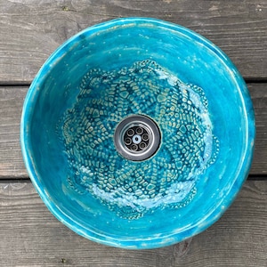 Puede incluir: Un lavabo de cerámica turquesa con un diseño estampado blanco y azul. El lavabo es redondo y tiene un desagüe en el centro.