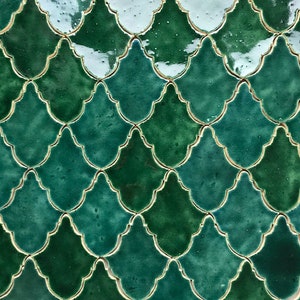 Pode incluir: Close-up de um padrão repetitivo de azulejos de cerâmica verdes. Os azulejos têm a forma de um quadrifólio estilizado com um acabamento brilhante e contornos dourados. A paleta de cores inclui tons de esmeralda e verde-azulado.