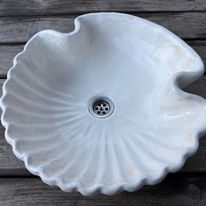 Peut inclure: Un évier blanc en forme de coquillage avec un bord festonné décoratif. L'évier a un drain central et est fait d'un matériau lisse et solide. L'évier est conçu pour être monté sur un comptoir et est un accessoire de salle de bain unique.