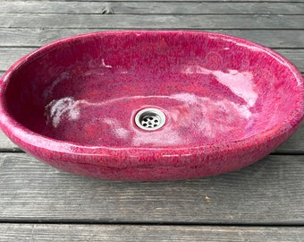 Puede incluir: Un lavabo de cerámica ovalado de color rojo oscuro con un desagüe plateado.