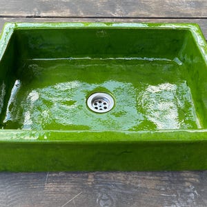 May include: A rectangular, glossy, lime green ceramic sink with a drain in the centre. The sink has a smooth, reflective surface and is set against a dark wooden background. The sink's design is simple and modern.