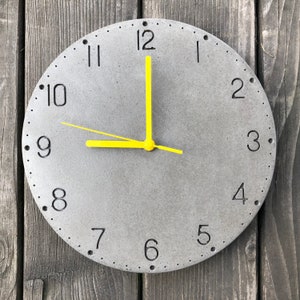 May include: A grey concrete clock with black numbers and a yellow hour and minute hand. The clock face has a subtle textured finish.