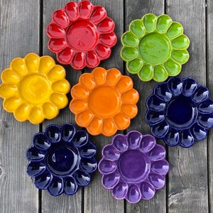 May include: Seven ceramic egg plates in different colors, including red, yellow, orange, green, blue, and purple. Each plate has a flower-shaped design with 12 individual indentations for holding eggs.