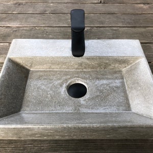Peut inclure: Un évier rectangulaire en béton avec un robinet et un drain noirs. L'évier a une surface texturée et un bord légèrement arrondi.
