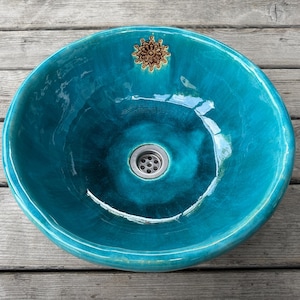 Könnte beinhalten: Ein rundes, türkisfarbenes Keramik-Aufsatzwaschbecken mit glänzender, nuancierter Oberfläche. Das Becken hat ein kleines, detailliertes braunes Mandala-Emblem am oberen Rand und einen mittigen, silberfarbenen Metallablauf am Boden.