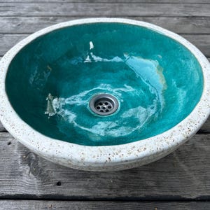 May include: A teal ceramic sink with a white rim and a drain in the center. The sink is sitting on a wooden surface.