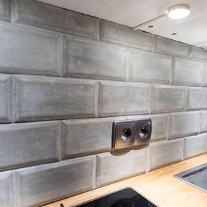 Könnte beinhalten: Nahaufnahme einer Küchenarbeitsplatte mit grauer Betonfliesen-Rückwand, einem Spülbecken aus Edelstahl und einem Wasserhahn. An der Rückwand befinden sich zwei Steckdosen.
