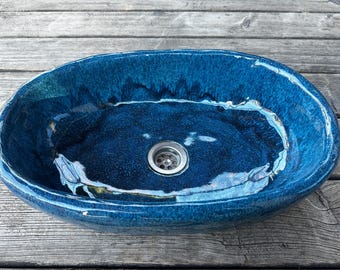 May include: Oval ceramic sink in a deep blue glaze. The sink features a textured surface and a metallic drain. The colour transitions from dark to light blue, creating a unique visual effect. A modern bathroom fixture.