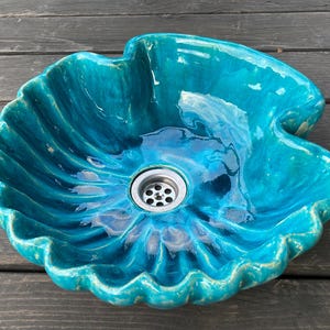 Könnte beinhalten: Ein glänzendes, türkisfarbenes Keramik-Aufsatzwaschbecken in Form einer großen, gewellten Muschel. Das Becken hat einen tiefen, geschwungenen Rand mit einer glatten, reflektierenden Glasur und einen mittigen, runden Edelstahl-Ablauf mit Sieb.