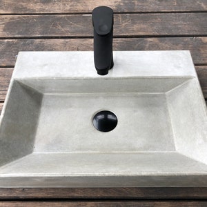 Könnte beinhalten: Ein rechteckiges Waschbecken aus Beton mit mattgrauer Oberfläche. Das Waschbecken hat einen schwarzen Wasserhahn und Abfluss. Das Waschbecken steht auf einer Holzoberfläche.
