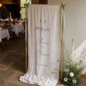 Könnte beinhalten: Ein hohes Hochzeits-Willkommensschild aus cremefarbenem Stoff, das an einem goldfarbenen Metallrahmen hängt. Das Schild zeigt die Initialen J E, den Text Welcome to the wedding of James & Eleanor und das Datum April 18, 2026 in schwarzer Schrift.