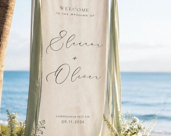 May include: A vertical off-white fabric wedding welcome sign suspended from a horizontal rod. The text reads: WELCOME TO THE WEDDING OF Eleanor + Oliver, SANDRINGHAM PE33 6IN, 09.11.2026. Sage green fabric ribbons are tied to the rod ends.