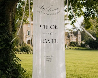 May include: A long, white, semi-sheer fabric banner hanging from a tree branch with white ribbon ties. The text reads: Welcome to the wedding of Chloe and Daniel, Chateau Rose, Provence, July 20th, 2026. The banner features a simple, elegant design.