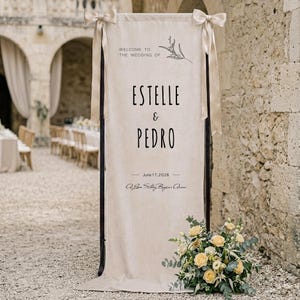 May include: A tall, rectangular beige fabric wedding sign suspended from a dark frame with light beige ribbons tied at the top corners. The text reads: WELCOME TO THE WEDDING OF ESTELLE & PEDRO, June 17, 2026, A Love Story Begins Anew.