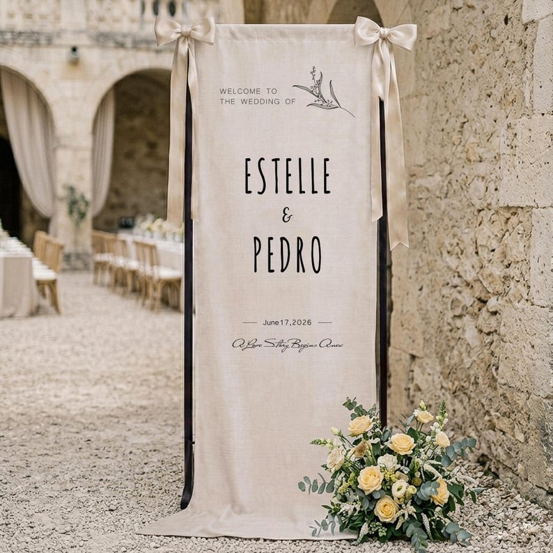 May include: A tall, rectangular beige fabric wedding sign suspended from a dark frame with light beige ribbons tied at the top corners. The text reads: WELCOME TO THE WEDDING OF ESTELLE & PEDRO, June 17, 2026, A Love Story Begins Anew.