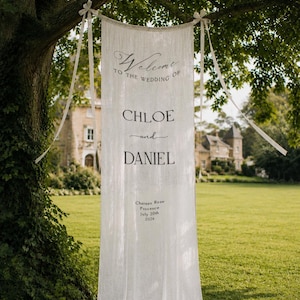 May include: A long, white, vertical fabric banner suspended from a tree branch with ribbons. The text reads: Welcome to the wedding of Chloe and Daniel, Chateau Rose, Provence, July 20th, 2026. The banner features a semi-sheer, textured material.