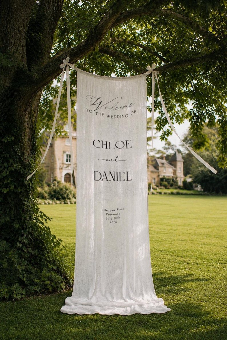 May include: A long, white, vertical fabric banner suspended from a tree branch with ribbons. The text reads: Welcome to the wedding of Chloe and Daniel, Chateau Rose, Provence, July 20th, 2026. The banner features a semi-sheer, textured material.