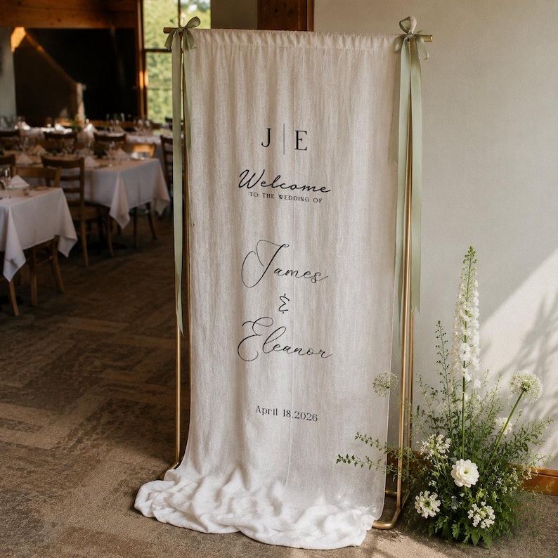 May include: A tall, off-white fabric wedding welcome sign hanging from a gold metal frame with sage green ribbons. The text reads: J E, Welcome to the wedding of, James & Eleanor, April 18, 2026. The fabric drapes onto the floor at the base.