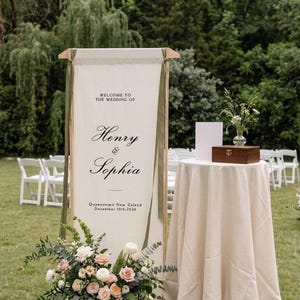 May include: A tall, rectangular fabric wedding welcome sign suspended from a wooden frame, featuring the text 'WELCOME TO THE WEDDING OF Henry & Sophia Queenstown New Zeland December 10th, 2026' with olive green ribbons and a floral arrangement.