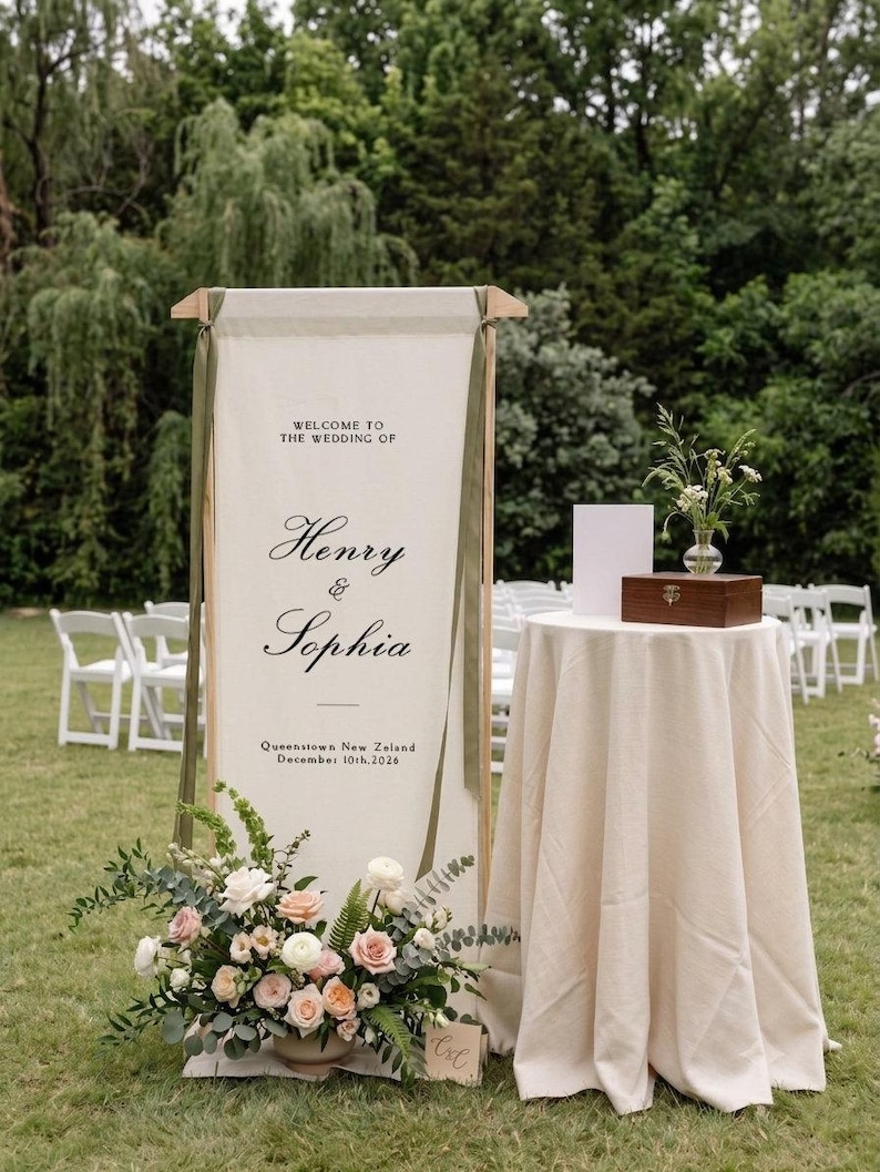 May include: A tall, rectangular fabric wedding welcome sign suspended from a wooden frame, featuring the text 'WELCOME TO THE WEDDING OF Henry & Sophia Queenstown New Zeland December 10th, 2026' with olive green ribbons and a floral arrangement.
