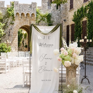 May include: A tall white fabric wedding welcome sign hanging on a dark metal frame, featuring the text J E, Welcome to the wedding of James & Eleanor, and April 18, 2026. The sign is draped with olive green velvet ribbon at the top.