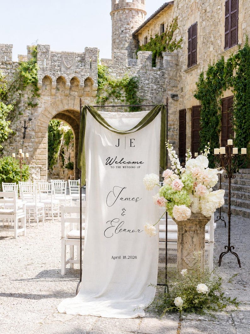 May include: A tall white fabric wedding welcome sign hanging on a dark metal frame, featuring the text J E, Welcome to the wedding of James & Eleanor, and April 18, 2026. The sign is draped with olive green velvet ribbon at the top.