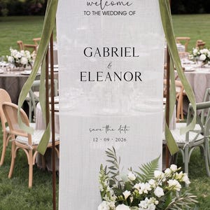 May include: A vertical white fabric wedding welcome sign suspended on a copper frame with olive green velvet ribbons. The text reads: welcome TO THE WEDDING OF GABRIEL & ELEANOR save the date 12 09 2026. A floral arrangement of white flowers sits at the base.