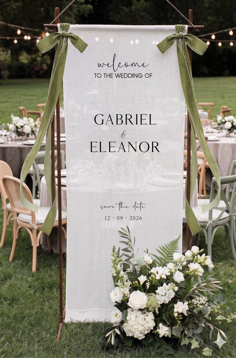 May include: A vertical white fabric wedding welcome sign suspended on a copper frame with olive green velvet ribbons. The text reads: welcome TO THE WEDDING OF GABRIEL & ELEANOR save the date 12 09 2026. A floral arrangement of white flowers sits at the base.