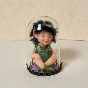 Könnte beinhalten: Eine kleine, grün gekleidete Feenfigur mit schwarzen Haaren und blauen Augen sitzt in einer Glaskuppel mit einem violetten Sockel. Die Kuppel steht auf einer weißen Oberfläche.