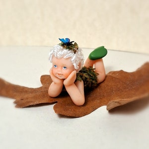 Könnte beinhalten: Eine kleine Porzellan-Feenpuppe mit weißen Haaren, blauen Augen und einem grünen, moosbewachsenen Kleid. Die Fee liegt auf einem braunen Blatt mit einem blauen Schmetterling auf dem Kopf.
