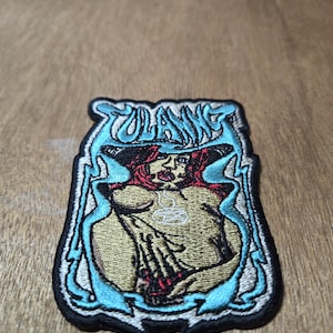 Pode incluir: Patch bordado com uma mulher de cabelo ruivo e um pingente de pentagrama. O patch tem uma borda azul e branca com o nome "Zolanne" no topo. O design está em um fundo preto.