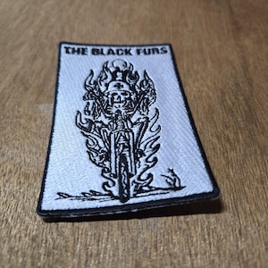 May include: A rectangular patch with a black border and a white background. The text "THE BLACK FURS" is at the top. A black and white graphic of a skull riding a motorcycle with flames is in the center.