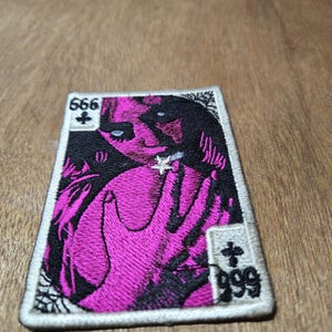 May include: Embroidered patch featuring a stylized image in pink and black. The design includes a figure with outstretched hands, the numbers 666 and 999, and a cross symbol. The patch has a white border.