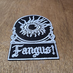 May include: Black and white embroidered patch with the word "Fangus!" in a stylized font. The patch features an eye design with sun-like rays above the text, and decorative borders.