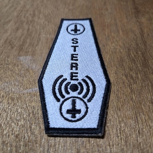 Pode incluir: Um patch em forma de caixão azul claro com detalhes bordados em preto. A palavra "STEREO" está centralizada verticalmente, com um sinal de mais em um círculo na parte superior e inferior, e um símbolo de onda de rádio no meio.