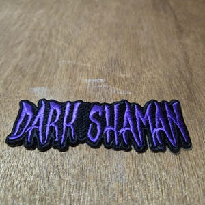 May include: A black embroidered patch with the words "DARK SHAMAN" in purple, stylized lettering. The patch has a black border and is likely designed to be sewn or ironed onto clothing or accessories.